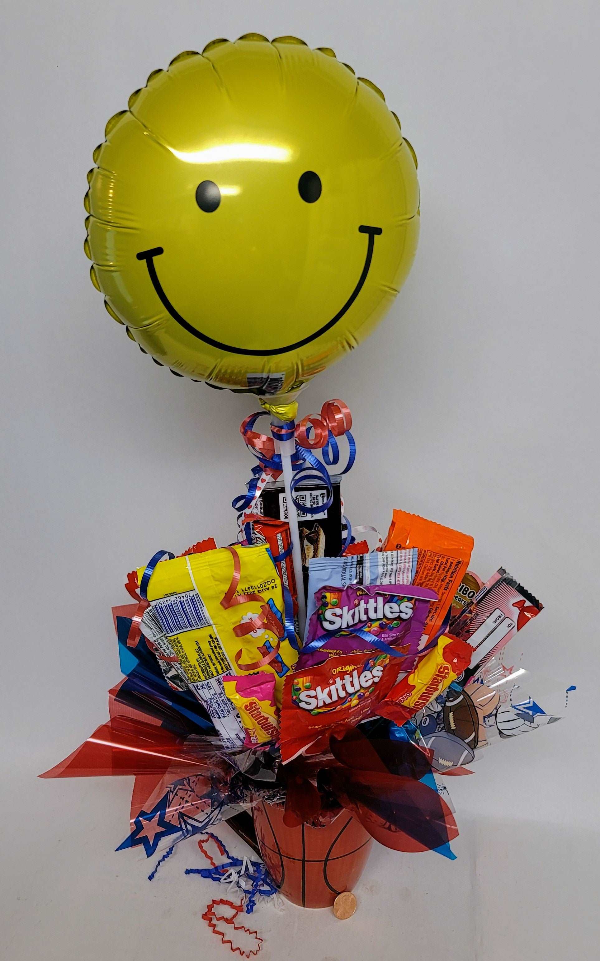 Assortment of candies bouquet in a Basketball ceramic mug with colorful ribbons, decorations and optional smiley face balloon.